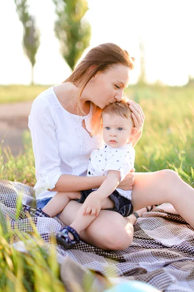 若い母親は、格子縞、背景の草の小さな子供のキス. — ストック写真