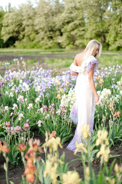 Young blonde girl wearing white dress standing near irises on garden. — Stock Photo, Image