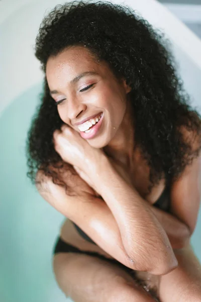 Photo rapprochée de jeune fille afro américaine heureuse assise dans le bain, portant un maillot de bain noir . — Photo