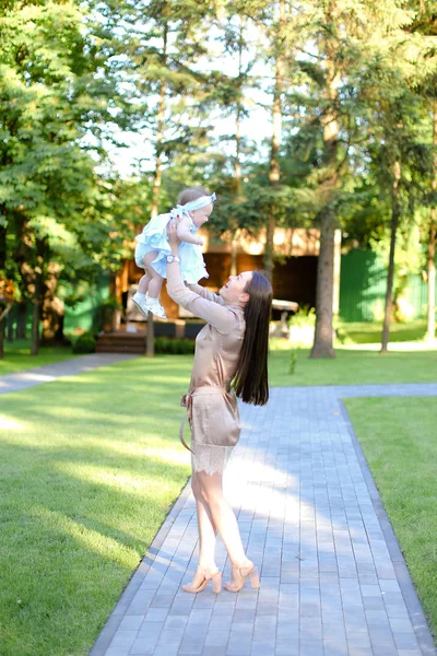 Jovem morena vestindo vestido segurando pequeno bebê no jardim . — Fotografia de Stock