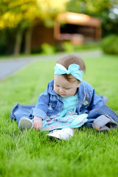 Pequeña niña caucásica sentada sobre hierba verde y con chaqueta vaquera . — Foto de Stock