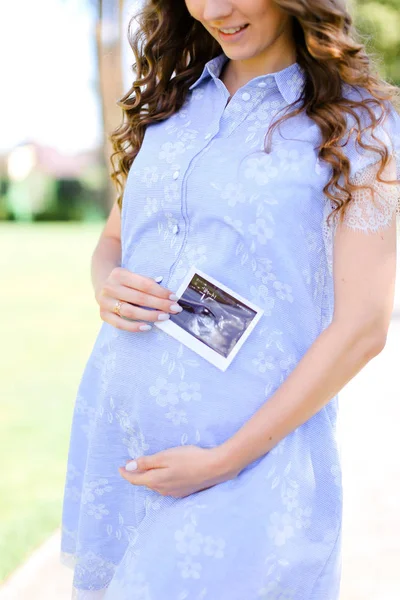 Feliz jovem grávida vestindo vestido azul segurando barriga com ultra-som . — Fotografia de Stock