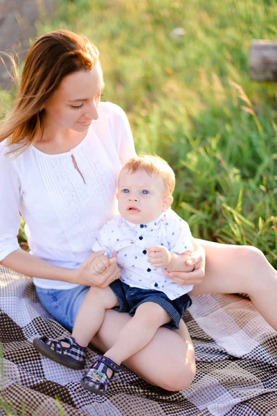 Ung glad mamma sitter med litet barn på pläd, gräs på bakgrunden. — Stockfoto