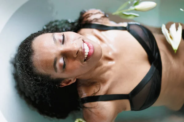 Young afro american relaxed woman taking bath and wearing black underwear, blue water background.