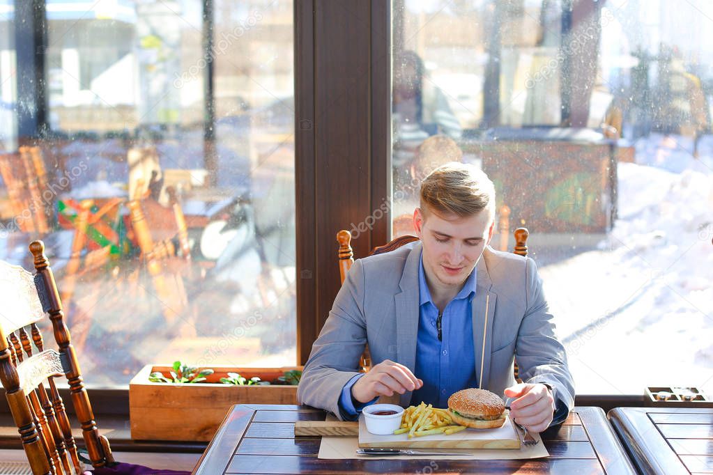 Portrait of busy young business man sitting with glass of drink and hamburger in fast food restaurant interior. Attractive caucasian man eating and using notebook computer in cafe. Indoors