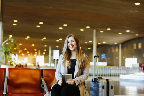 Młoda kobieta uśmiechający się siedząc przy hali lotniska z tabletu i walizka. — Zdjęcie stockowe