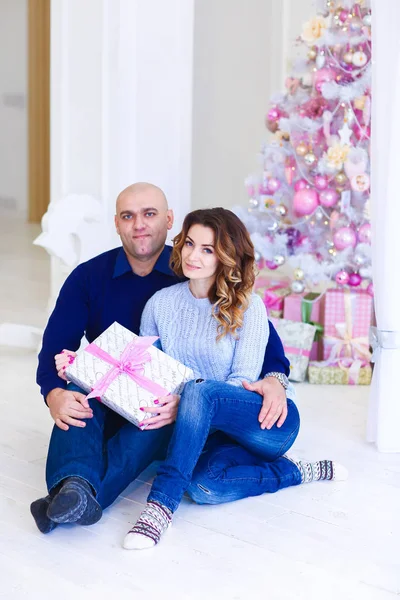 Junges glückliches Paar sitzt neben geschmückter rosa Tanne mit Geschenk. — Stockfoto