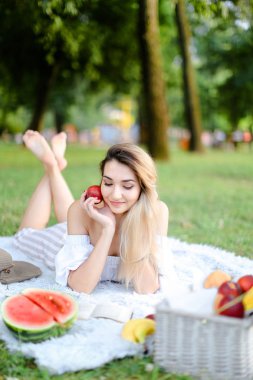 Parkta ekoseye uzanmış, karpuz yanında kitap okuyan ve elma saklayan genç bir kadın..