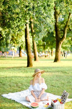 Bahçede ekoseli meyvelerin yanında oturan şapkalı genç bir kadın, arka planda çimenler..