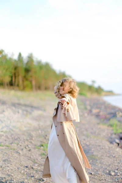 Young woman wearing coat and white dress standing on sand with trees in background. — Stock Photo, Image
