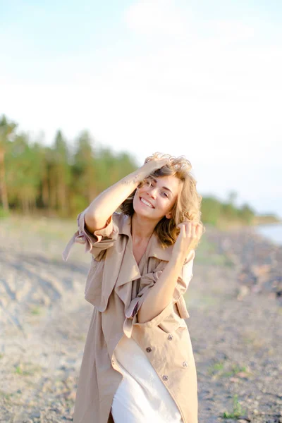 Young caucasian girl wearing coat and white dress standing on sand with trees in background. — Stock Photo, Image