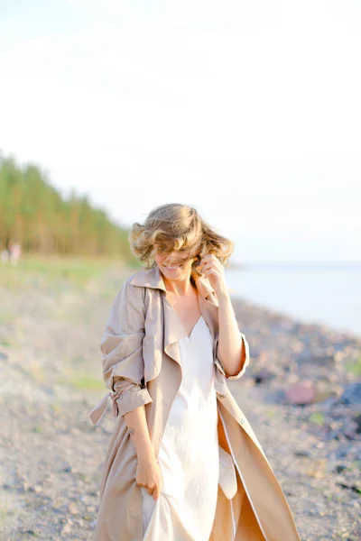 Giovane donna sorridente in piedi sulla spiaggia e indossa cappotto con abito bianco . — Foto Stock