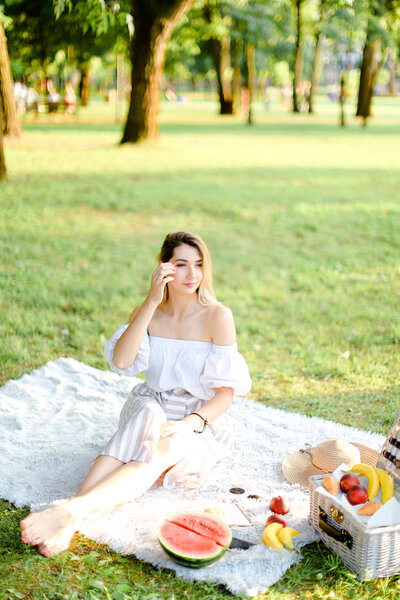 Young woman having picnic on plaid and sitting in park with fruits.