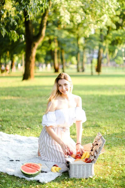 Молодая счастливая девушка готовится к пикнику в парке, сидит на клетке рядом с коробкой и ватерлимон . — стоковое фото