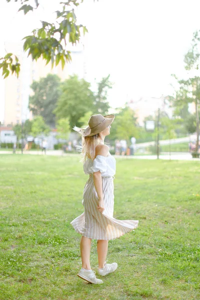 Back view of young girl wearing hat and dress, standing in park. — Stock Photo, Image