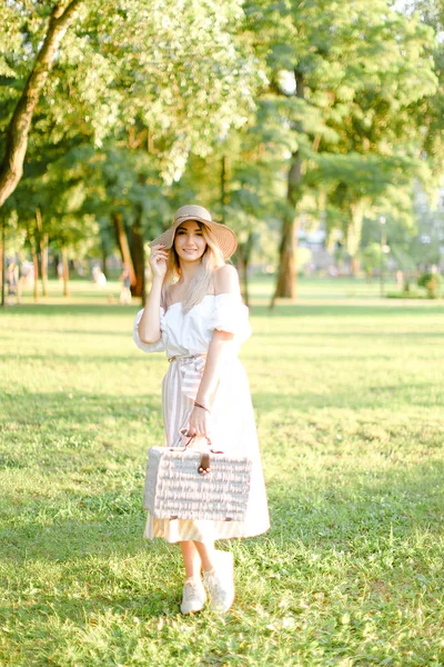 Young blonde girl wearing hat standing in park with bag. — ストック写真