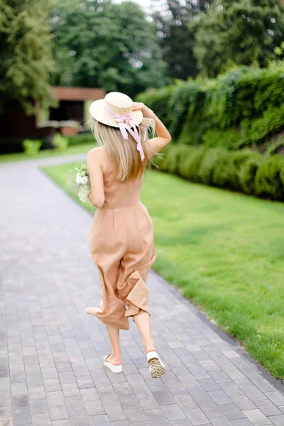 Back view of young blonde female person in body color overalls and hat with flowers. — Stock Photo, Image