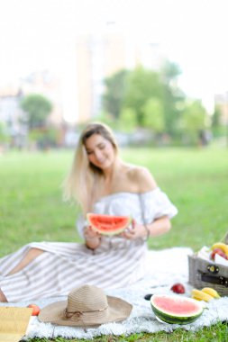 Meyve ve şapkanın yanında ekose üzerinde oturan, karpuz yiyen, arka planda çimen yiyen genç bir kadın..