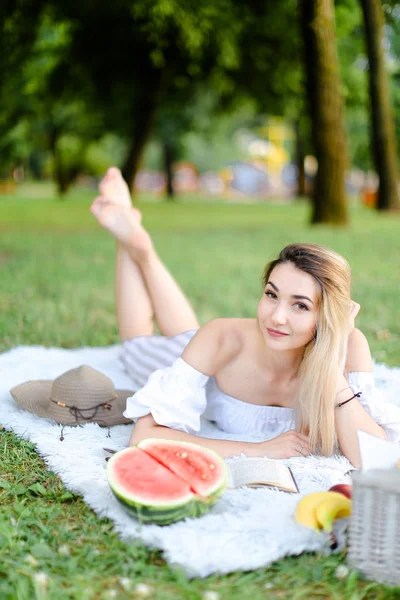 Junge Frau liegt auf Plaid im Park, liest Buch in der Nähe von Wassermelone und Hut. — Stockfoto