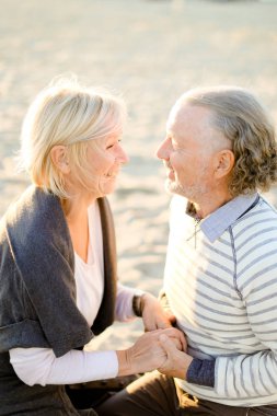 Yaşlı gülümseyen karısı el ele tutuşarak kıdemli koca ve kum plajda oturan.