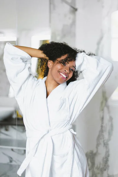 Young afro american nice woman standing in bathroom and wearing white bathrobe.