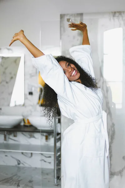 Young afro american woman standing in bathroom and wearing white bathrobe. — Stock Photo, Image