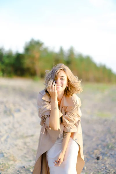 Sorridente giovane donna che indossa cappotto in piedi sulla sabbia con alberi in background . — Foto Stock