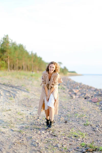 Smiling young woman standing on sea beach and wearing coat with white dress. — Stock Photo, Image