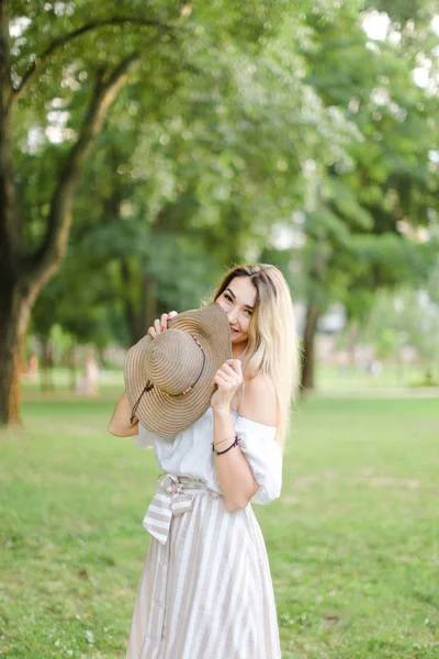 Young jocund woman standing in park and keeping hat. — Stock Photo, Image