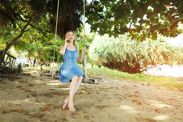 Zonnige lichte portret van de jonge blonde meisje praten door smartphone en rijden schommel in Bali, zand in de achtergrond. — Stockfoto