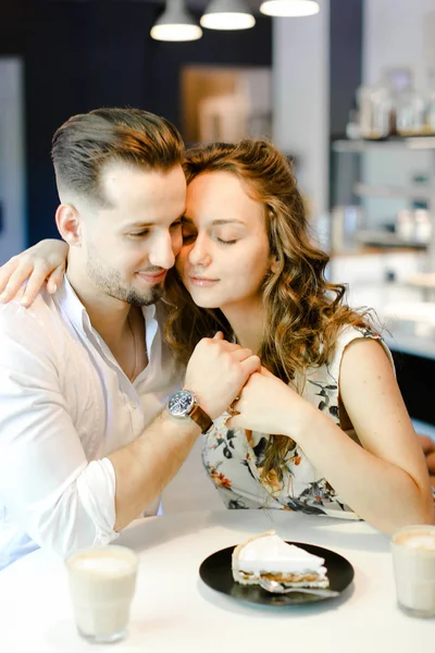 Joven linda pareja sentada en la cafetería con tazas de café, pastel y abrazos . — Foto de Stock