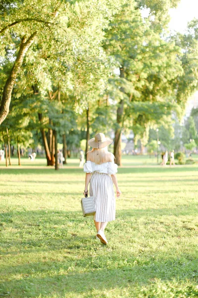 Vista posteriore della giovane donna americana in cappello che cammina nel parco e tiene borsa e occhiali da sole . — Foto Stock