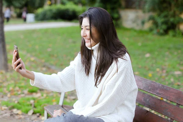 Chica japonesa sentada en el banco y haciendo videollamada por teléfono inteligente . — Foto de Stock