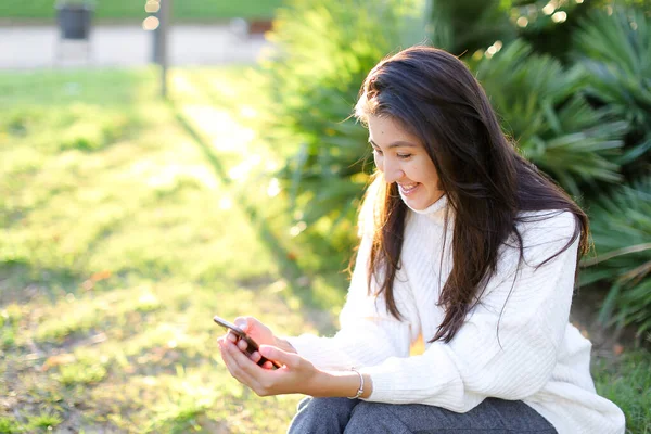 Chica asiática en suéter blanco sentado en el parque y el uso de teléfono inteligente . — Foto de Stock