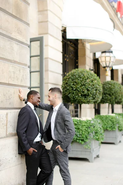 Afro-americano e caucasiano feliz gays em ternos de pé perto de construção e falando fora . — Fotografia de Stock