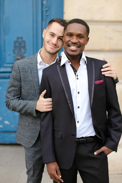 Двоє усміхнених чоловіків, кавказький і афроамериканський, одягнені в костюми, що стоять біля будівлі і обіймаються . — стокове фото
