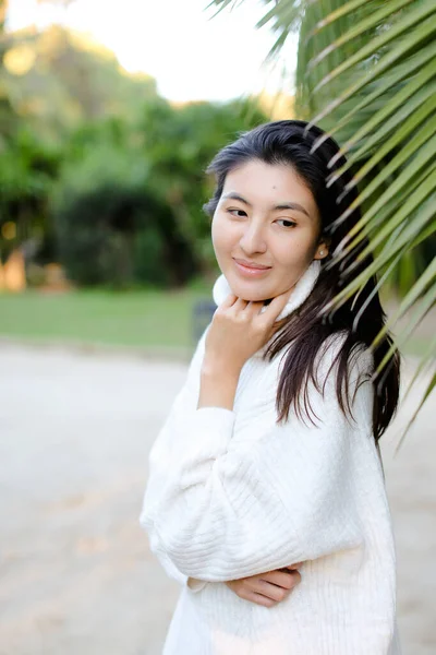 Chinese nice girl in white sweater standing near palm leaf. — Zdjęcie stockowe