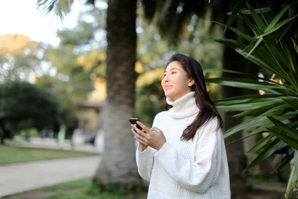 Chino buena chica usando teléfono inteligente y caminando en el parque tropical . — Foto de Stock