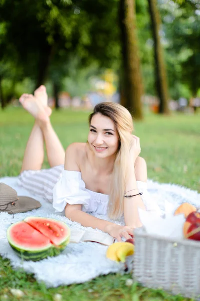 Jeune belle femme couchée sur plaid dans le parc, livre de lecture près de pastèque et chapeau . — Photo