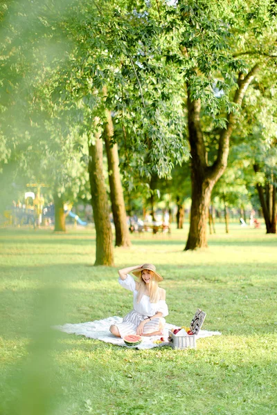 Giovane donna bionda caucasica in cappello appoggiata su plaid con frutta nel parco . — Foto Stock