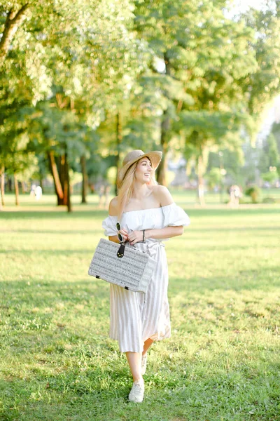 Ung söt kvinna i hatt promenader i parken och hålla väska och solglasögon. — Stockfoto