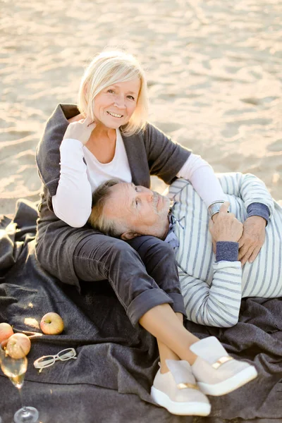 Esposo sênior deitado de joelhos esposa e xadrez, champanhe e frutas na praia de areia . — Fotografia de Stock