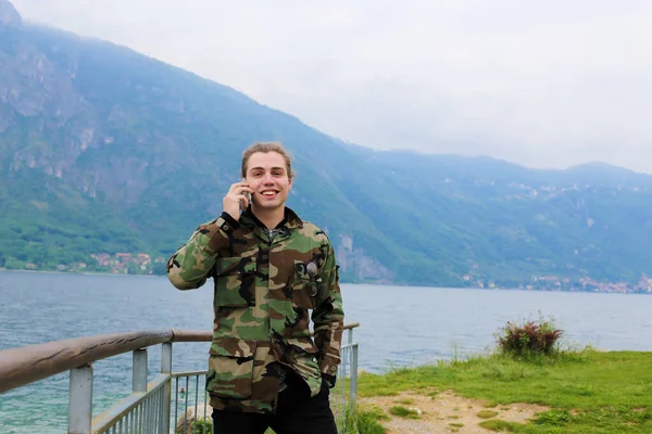 Hombre caucásico hablando por teléfono inteligente cerca de barandilla, lago Como y la montaña de los Alpes en el fondo . — Foto de Stock