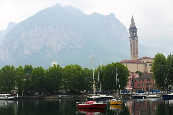 Barcos no lago Como perto da igreja com torre, árvores e montanha no fundo . — Fotografia de Stock