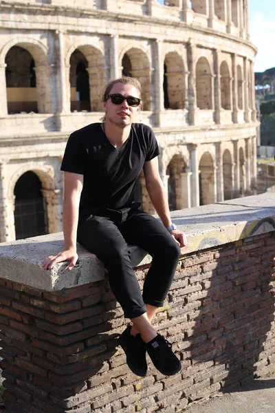 Fiatal kaukázusi Férfi turista szemüveg ül a téglafal Colosseum háttérben, Róma, Olaszország. — Stock Fotó