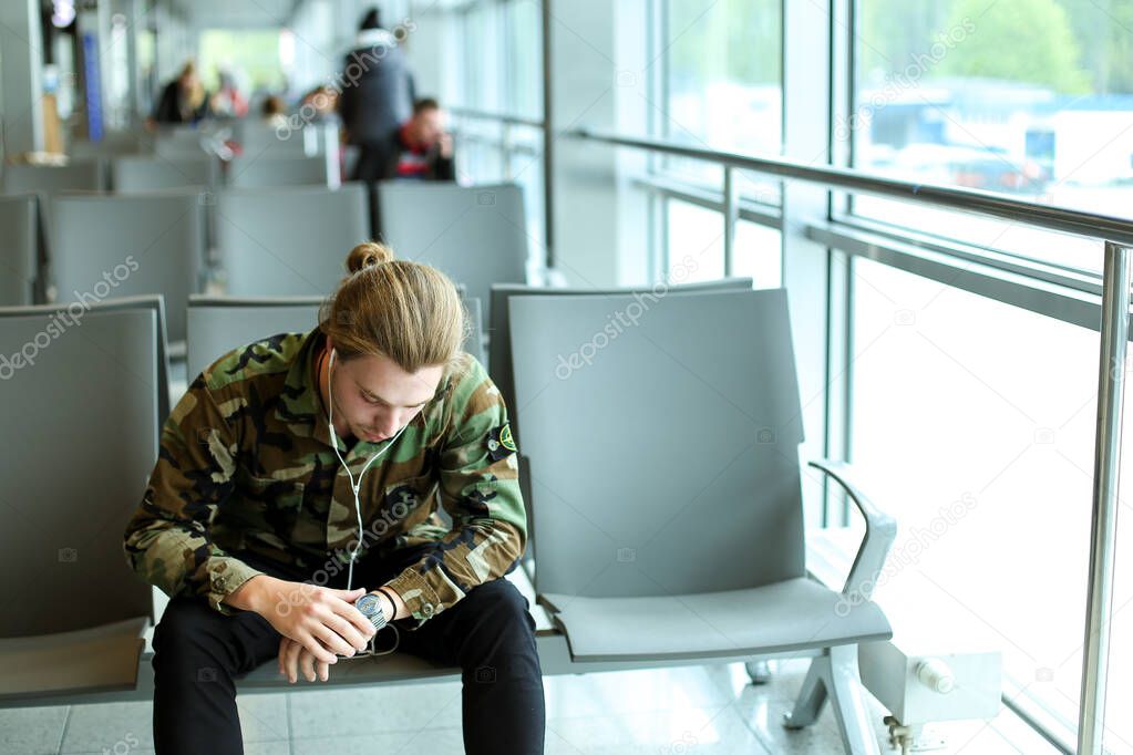 Young man sitting in waiting hall at airport, listening to music with earphones and looking at watch.