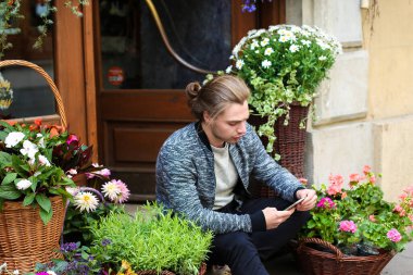 Genç erkek kişi tarafından akıllı telefon sohbet ve sepetler içinde buketleri yakınındaki çiçek dükkanında oturmuş.