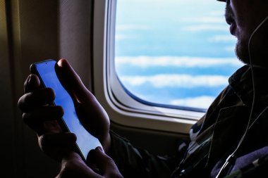Yakın plan sakallı adam uçağın penceresinin yanında akıllı telefon kullanıyor..
