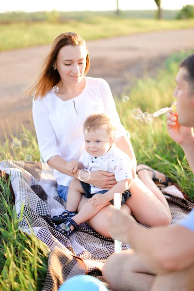 Giovani genitori caucasici felici seduti sull'erba con piccolo bambino e bolle che soffiano . — Foto Stock