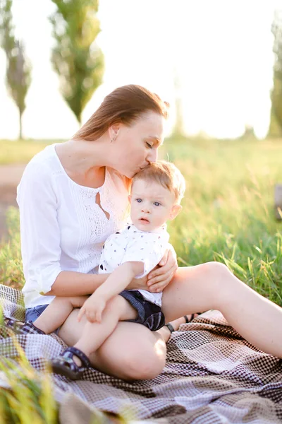 Giovane madre bionda seduta con bambino su plaid, erba su sfondo . — Foto Stock